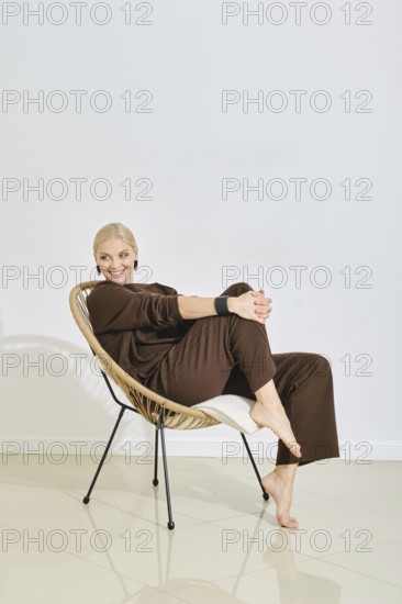 A barefoot woman dressed in brown lounge wear relaxes in a stylish chair, smiling and exuding a sense of calmness. Soft lighting highlights her content expression in a minimalistic indoor setting