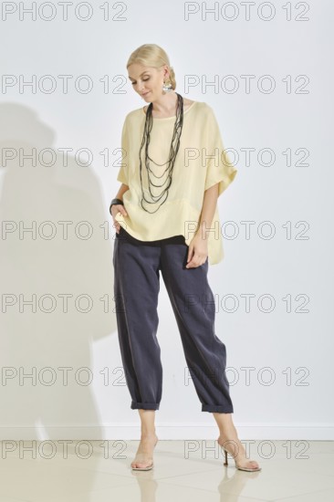 A woman stands confidently wearing a stylish yellow blouse and dark pants, accessorized with a necklace. Soft lighting highlights her relaxed pose and contemporary style