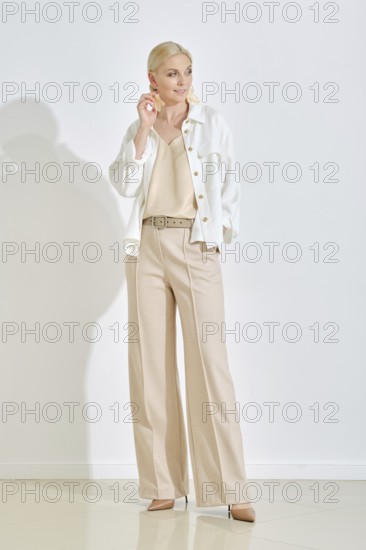 A woman dressed in a cream top and stylish beige trousers strikes a confident pose in a minimalist interior. She pairs the outfit with a light jacket, exuding elegance and sophistication