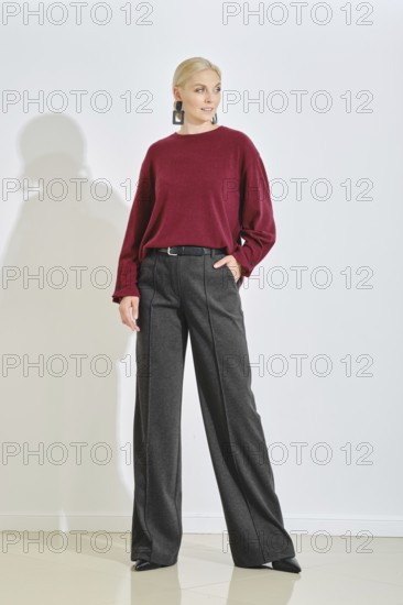 A fashion model stands confidently in a minimalist studio, wearing a maroon sweater and high-waisted black trousers. Bright daylight illuminates the space, enhancing her stylish look