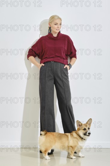 A woman dressed in a burgundy sweater and gray wide-leg pants stands confidently next to her corgi. The bright indoor space highlights her modern style while showcasing the playful dog beside her