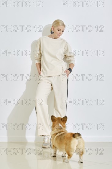 A woman dressed in light, casual attire stands against a plain wall while a corgi dog curiously looks up at her. The sunlight creates soft shadows, enhancing the warm atmosphere of the moment