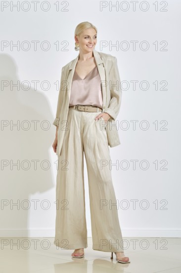 A confident woman stands in a well-lit indoor area, wearing casual outfit featuring a light-hued blazer and wide-leg trousers. Her expression conveys a sense of poise and sophistication