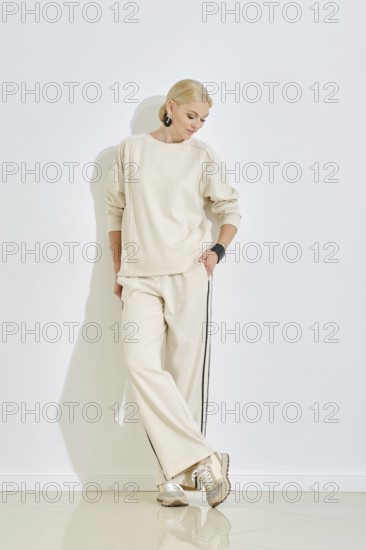 A woman dressed in a cozy beige outfit leans casually against a white wall, showcasing a relaxed yet stylish look. Her light blonde hair complements the minimalistic aesthetic of the setting
