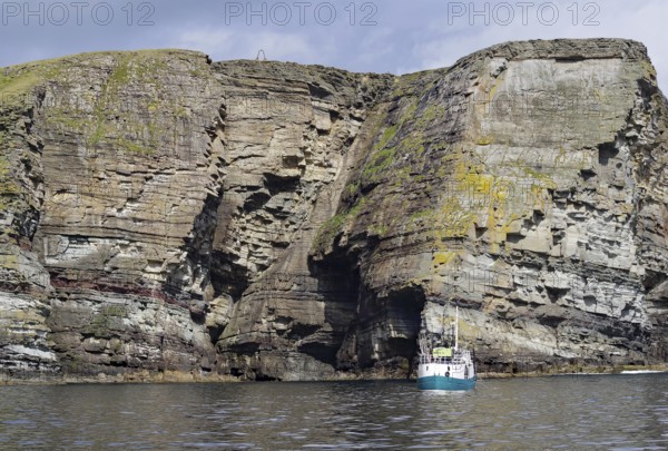 Boat in front of massive cliffs in the sea under a cloudy sky, Noss, Shetland Islands, Scotland, United Kingdom