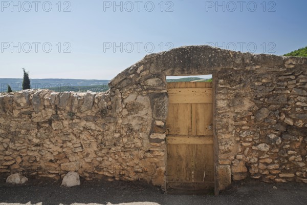Stone wall with wooden gate and view of a wide landscape under a blue sky, Provence, France