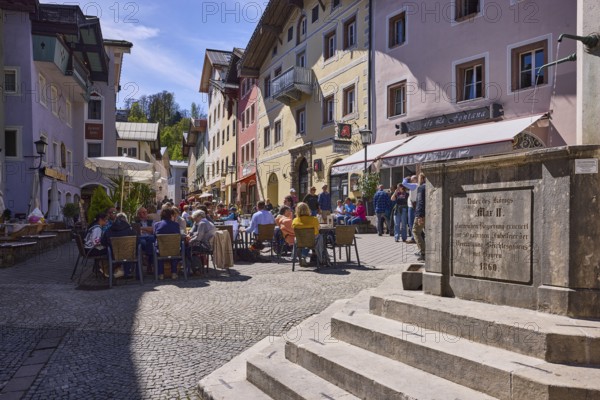 Market fountain, stone fountain, pedestrian zone, stairs, general architecture, historical residential and commercial buildings, outdoor area of a restaurant, pedestrians and guests as secondary motif, hill, trees, blue sky, cirrostratus clouds, market square, Berchtesgaden, Upper Bavaria, district Berchtesgadener Land, Bavaria, Germany