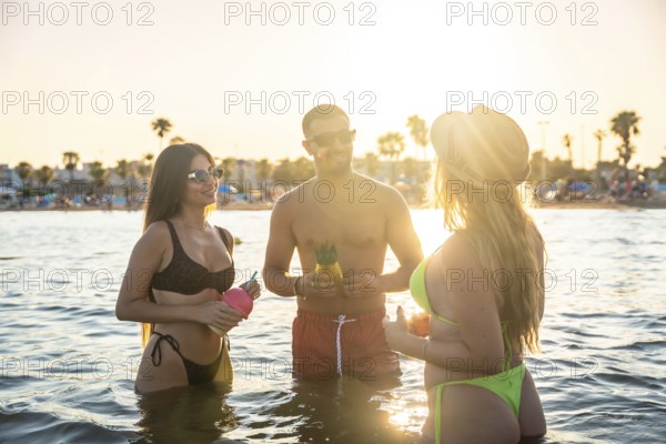 Three friends enjoying pineapple cocktails in the sea at sunset, celebrating summer vacation with laughter and joyful conversations