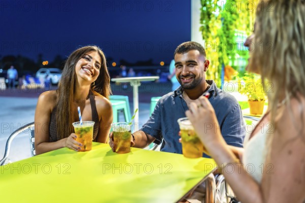 Three friends are laughing and chatting while enjoying refreshing beverages at a vibrant outdoor cafe on a warm summer evening
