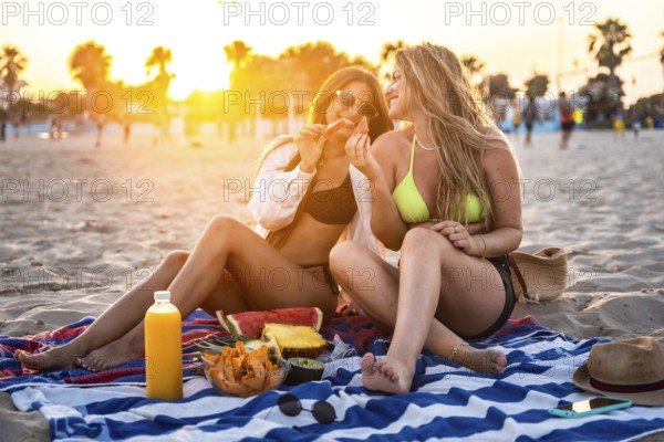 Two happy women sharing watermelon slices during a vibrant sunset picnic on the beach, enjoying snacks and drinks