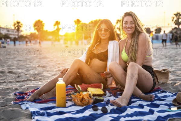 Two friends savoring a delightful picnic on the beach, surrounded by fresh fruit and juice, while enjoying a stunning sunset