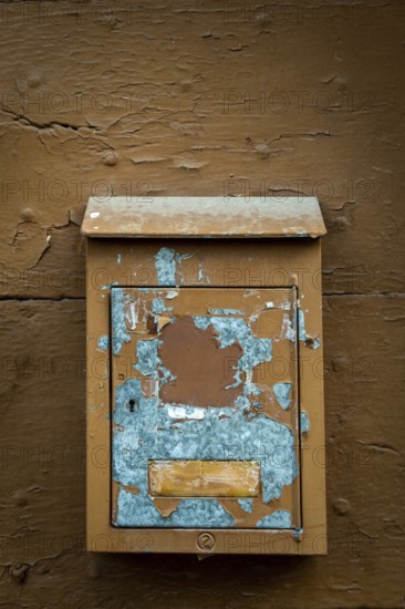 A rustic mailbox is mounted on a weathered brown wall, featuring peeling paint and faded colors. The blue paint reveals its age, while the wooden backdrop shows texture and wear. France