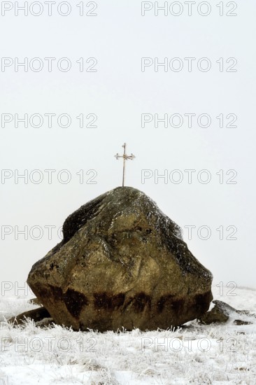 A large rock topped with a cross stands starkly against the winter landscape of the Aubrac Plateau in Lozere, Occitanie, France