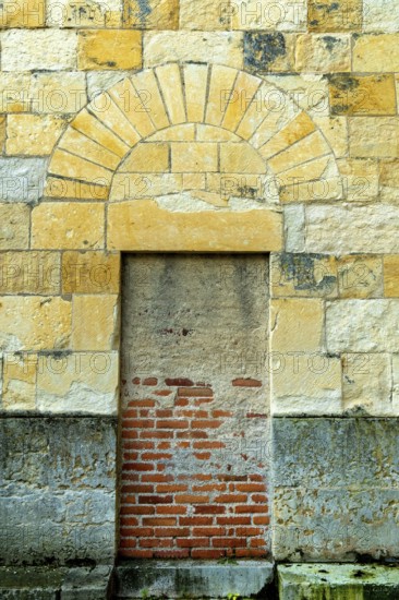An old stone wall showcases a prominently arched doorway, leading to an unseen area beyond. The lower brick section highlights the texture and age of the structure. France