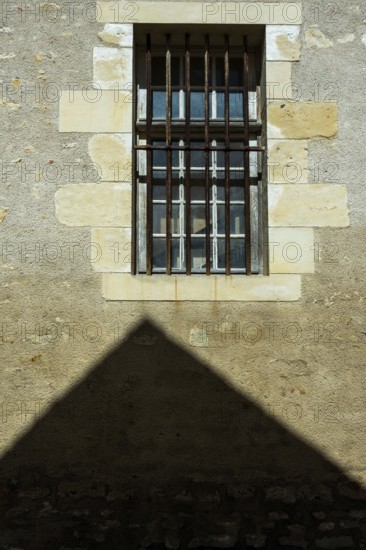 A barred window positioned on a rough stone wall casts a strong shadow below. Sunlight enhances the texture of the wall, creating a striking visual contrast during the day. France