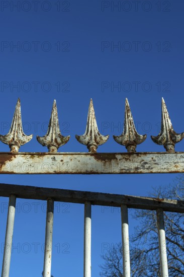 A weathered wrought iron fence stands tall under a bright blue sky. The fence has pointed spikes, showcasing signs of rust and age, adding character to the setting. France