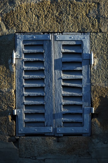 Sunlight casts shadows on a pair of weathered blue shutters attached to an old stone wall. The rustic details add character and charm to the architectural setting of the scene. France