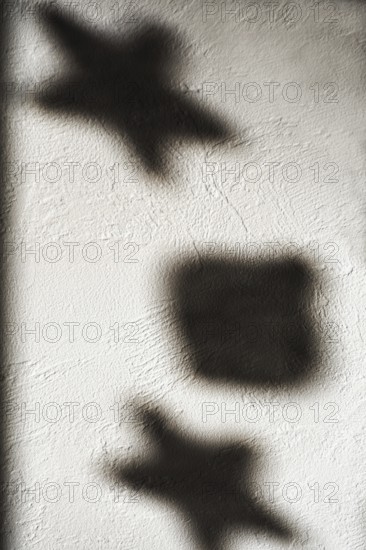 Shapes of stars and a square cast shadows on a textured white wall, illuminated by gentle light. The interplay of light and shadow creates a captivating visual composition