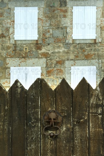 A weathered wooden fence stands in the foreground, with a charming stone house featuring white shutters in the background. Occitanie. France