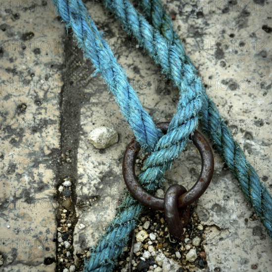 A close-up view reveals a vibrant blue rope intricately intertwined with a metal ring, resting on a textured and worn surface scattered with debris, highlighting the contrast in materials