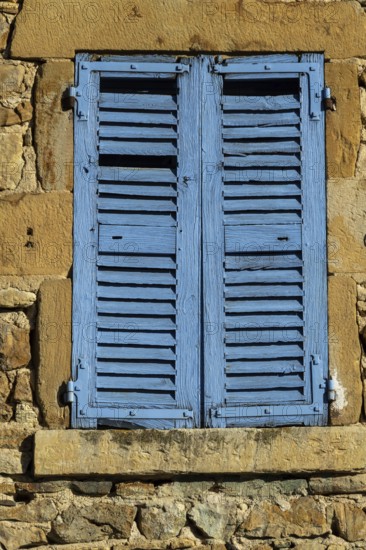 Bright blue shutters adorn the weathered stone wall of a traditional building in a picturesque village. Sunlight reflects off the vibrant color, creating a cheerful atmosphere. France