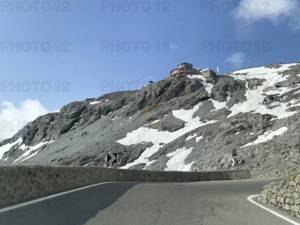 View in front of a narrow bend above the tree line in front of the Stelvio Alpine Pass road to the rocky mountain peak and the Hotel Tibethaus restaurant, Stelvio, Autonomous Province of Bolzano, South Tyrol, Italy