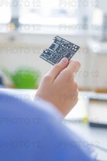 Hand holds a circuit board in front of a blurred background, window light shines in