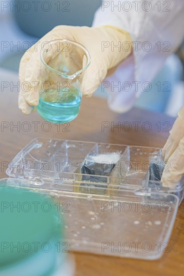 Person in gloves holding a beaker with blue liquid over a tub, Battery Research, Nagold, Germany