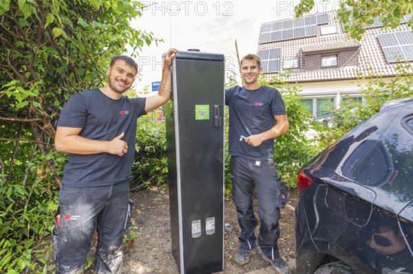 Two technicians pose proudly next to a closed power box outdoors, installing a charging station for electric cars, Calw, Germany