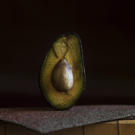 A half avocado is placed on a rough, textured surface, highlighting its smooth, creamy interior and prominent seed. The low lighting creates a rich, alluring atmosphere around the fruit