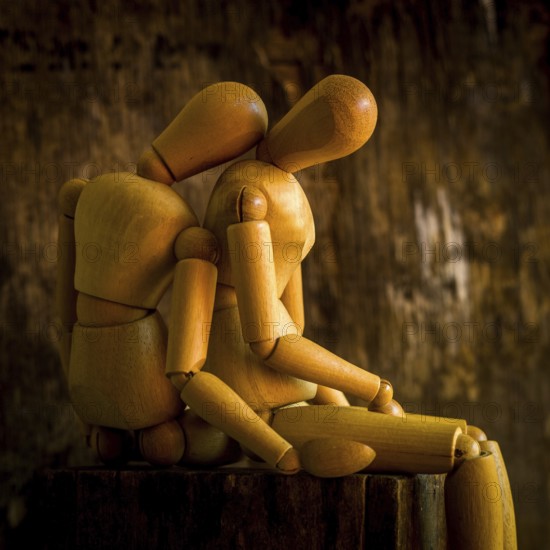 A wooden mannequin is positioned on a rough wooden surface, looking contemplative against a textured, dimly lit background. The warm tones enhance the somber mood of the scene