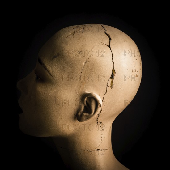 A head of a broken woman dummy is positioned in profile. The damaged surface reveals cracks and marks against a stark black background, emphasizing its worn features