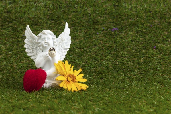 A white angel figure with a red heart and yellow flower on grass