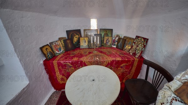 A table with red blankets and several icons flanked by candles in a small room, chapel, church, Agios Isidoros, rocky islet, Gourna Bay, Kokkali, Leros, Dodecanese, Greek Islands, Greece