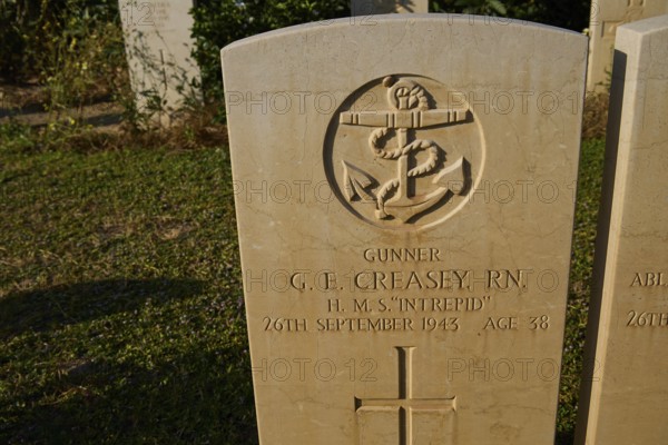 A maritime emblem on a gravestone with personal information, surrounded by grass and shadow, sinking of the H.M.S. Intrepid, military cemetery, Commonwealth War Cemetery, military cemetery, WW2, 1939-1945, 179 graves, Alinda, Leros, Dodecanese, Greek Islands, Greece