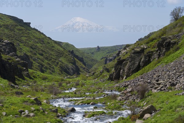 A river runs through a rocky landscape with a snow-covered mountain in the distance, Our Lady Church with Ararat in the background, Vahramashen Church, Vahramashen Surp Astvatsatsin, Amberd Gorge, Aragazotn Province, Armenia