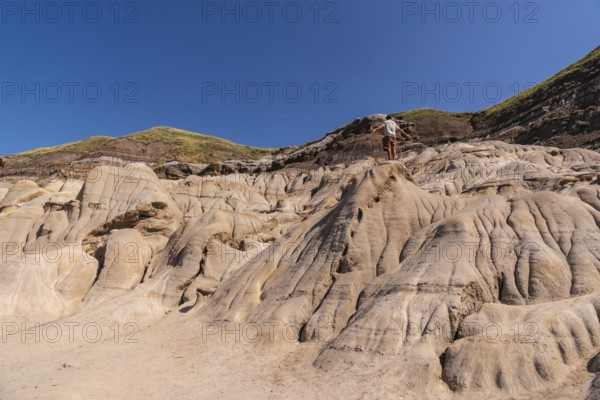 Tourist standing on a hoodoo rock formation, exploring the unique badlands landscape of drumheller, alberta, under a clear blue sky, enjoying the vastness and beauty of nature
