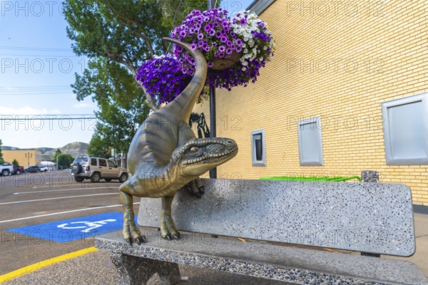 Dinosaur statue resting on a bench in front of a brick building, surrounded by flower baskets in drumheller, alberta, a town famed for its paleontological history