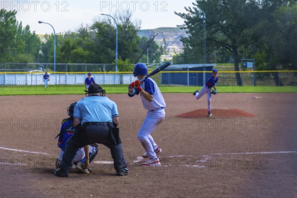 Teenage baseball batter waiting for the pitch while the pitcher is throwing the ball during a sunny summer game, with the umpire and the catcher behind him