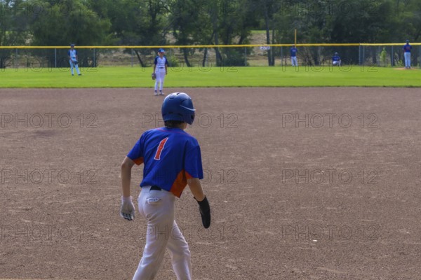 Young baseball player is walking on the infield during a game, wearing a blue and orange jersey with the number one, white pants, a blue helmet, and batting gloves