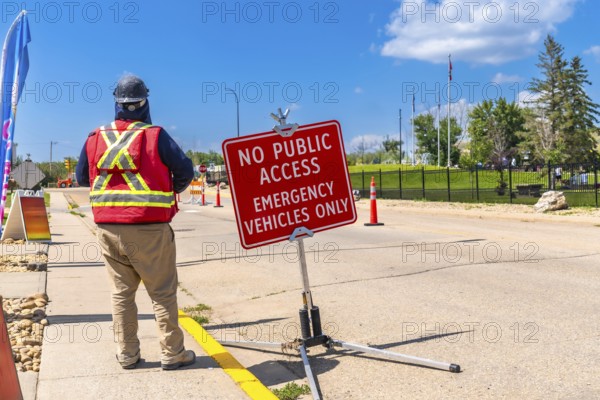 Construction worker wearing a safety vest and hardhat, standing near a no public access sign, controlling road access for emergency vehicles on a sunny day in drumheller, alberta