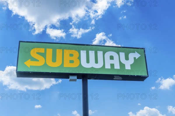 Green and yellow sandwich restaurant sign with white arrow pointing towards restaurant entrance against bright blue sky with fluffy white clouds, indicating location of popular fast food chain