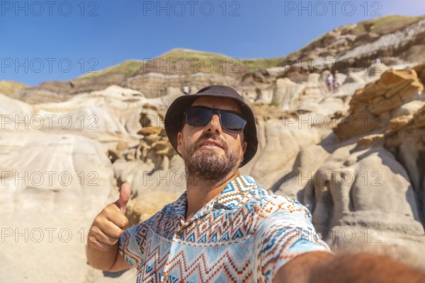 Happy tourist wearing sunglasses and a bucket hat giving a thumbs up while taking a selfie in front of unique sandstone hoodoos under a clear blue sky in drumheller, alberta
