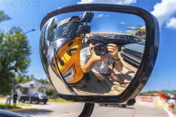 Photographer is taking a selfie reflected in the mirror of a truck, with a yellow school bus and parked cars in the background, in drumheller, alberta, on a sunny day
