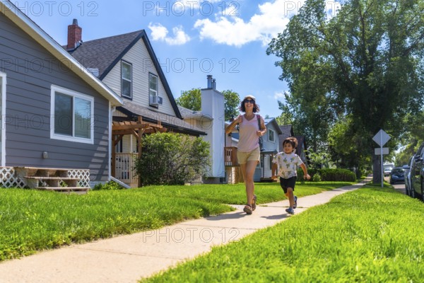 Mother and her young son are running on the sidewalk in a quiet, residential neighborhood on a sunny summer day, enjoying quality time together