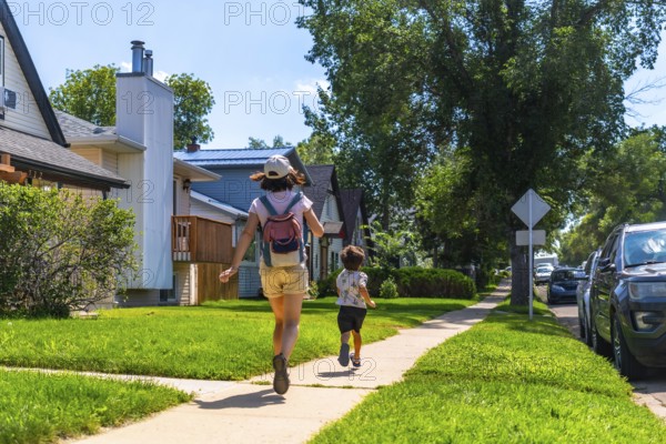 Mother with backpack and her son are running down a sidewalk in a residential area of drumheller, alberta, canada, during a sunny summer day, showcasing the peaceful suburban life