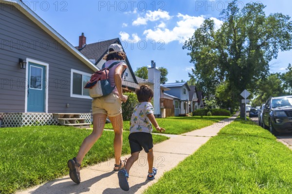 Mother with backpack and her son are running on the sidewalk in a residential area of drumheller, alberta, during a sunny summer day, showcasing the peaceful suburban life