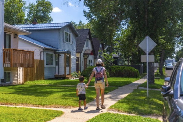 Mother and her young son are enjoying a leisurely stroll along a sidewalk in a quiet residential neighborhood, passing by charming houses with green lawns and trees on a sunny summer day