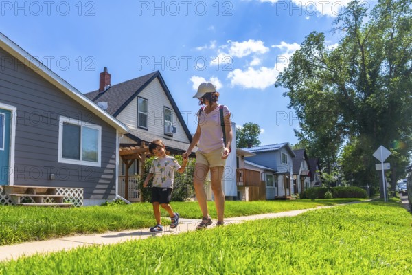 Mother and son walking hand in hand on a sunny day along a sidewalk in a peaceful drumheller neighborhood, surrounded by green lawns and charming homes