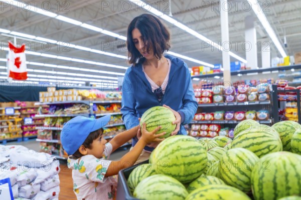 Mother and son selecting a large, ripe watermelon in a supermarket, enjoying quality time together while surrounded by an abundance of fresh produce in the vibrant grocery aisle